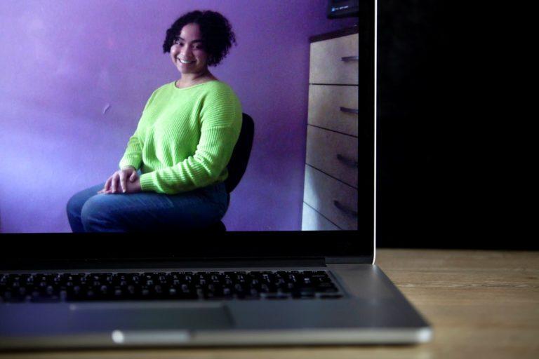 Sabrina Salas, sitting in a chair and wearing a green sweater, is shown on the screen of a laptop.