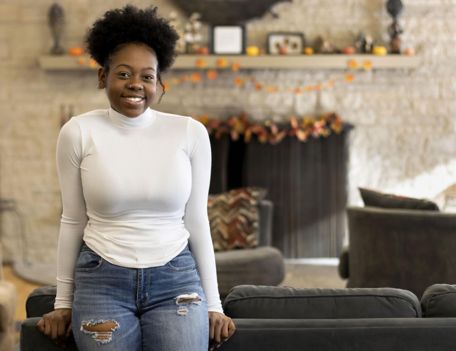 Adona Lauriano, wearing a white turtleneck and blue jeans, leans against a dark grey couch.