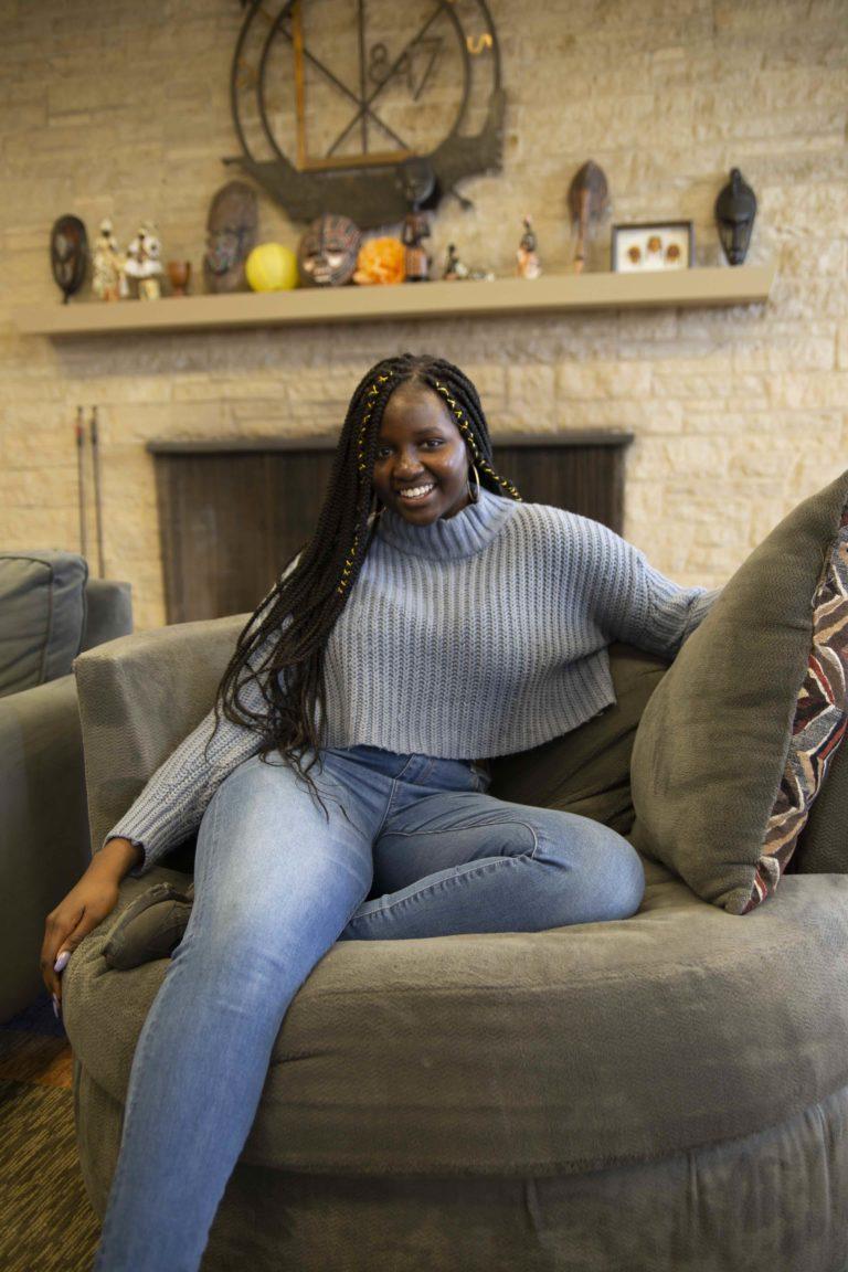 Awa Badiane, wearing a grey sweater and blue jeans, sits on a circular lounge chair and smiles at the camera.