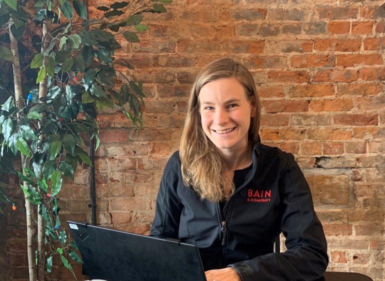 Emily Muhs, wearing a "BAIN" jacket, smiles at the camera against a brick wall.