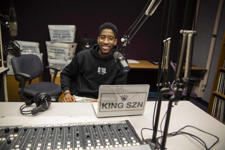 De Andre King, surrounded by DJ equipment, smiles at the camera.