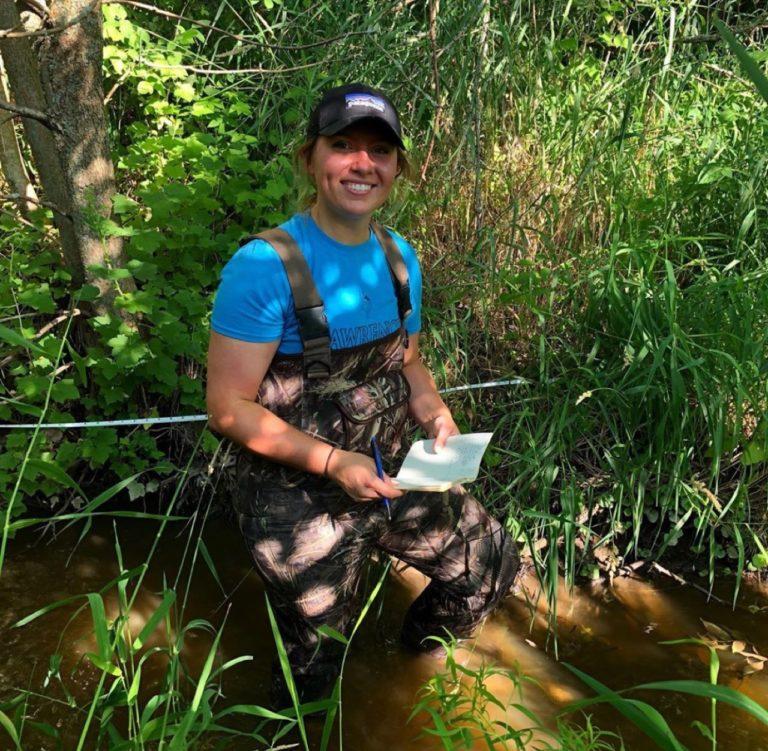 Summer Kopitzke squats in a creek, surrounded by greenery.