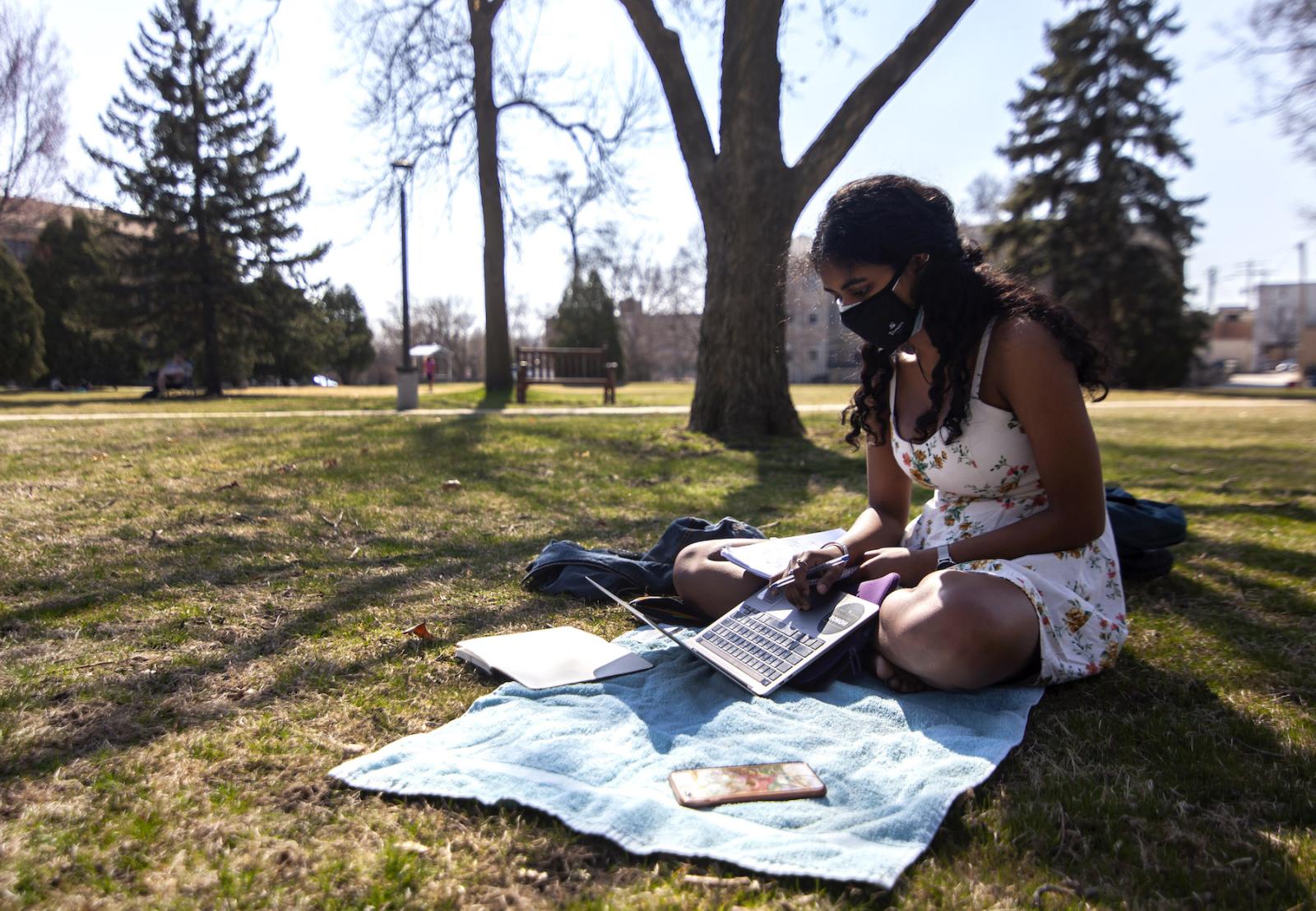 Adya '23 sitting on Main Hall Green reading with a mask on.