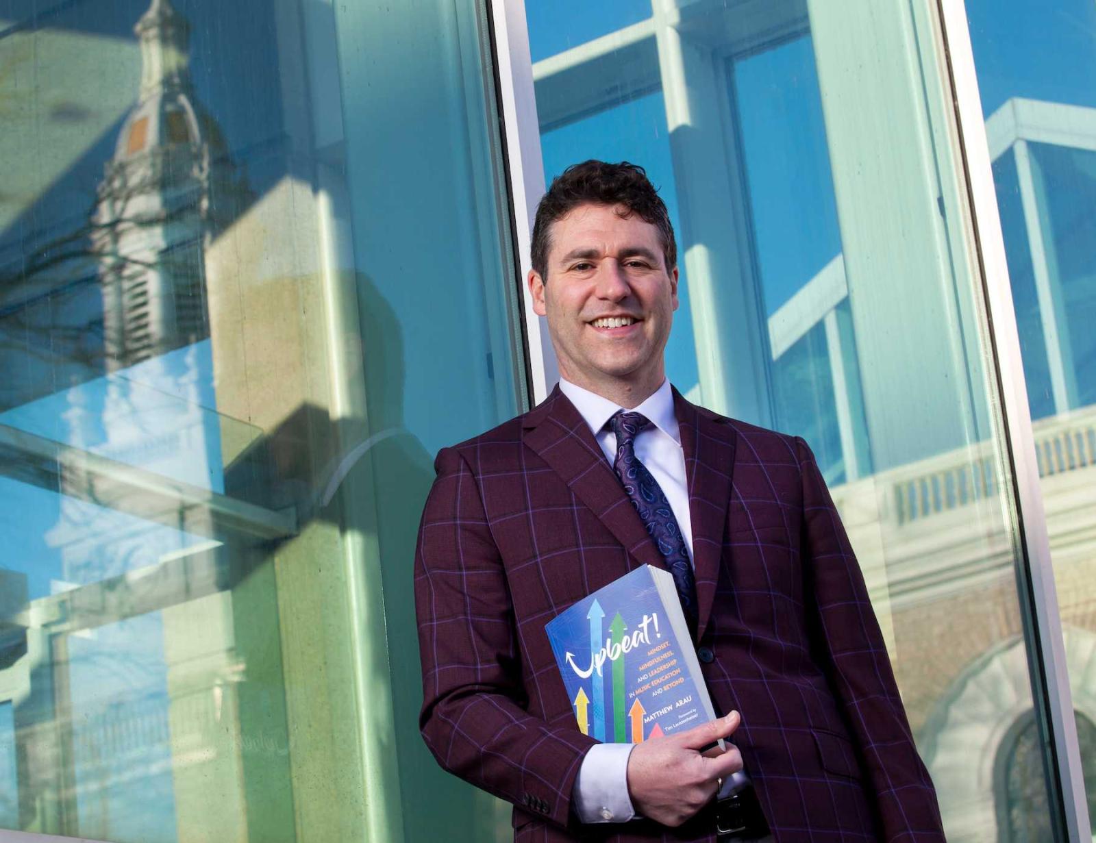 Matthew Arau holds his book, Upbeat!, as he stands near the Music-Drama Center with Memorial Chapel reflected in the windows.