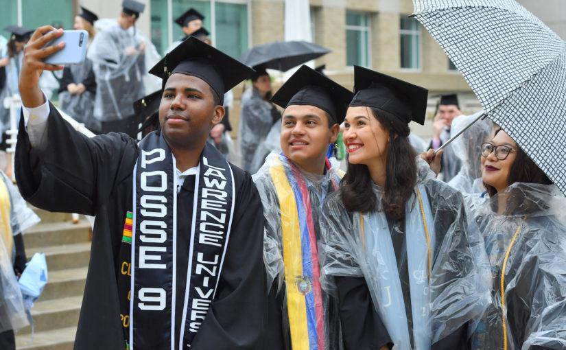 Three Lawrentians in black cap and gowns with clear plastic rain ponchos smile for a selfie.