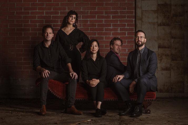 Five people sit and pose on a red velvet couch against a brick wall, dressed in dark formal clothing, looking at the camera.