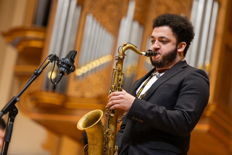 A musician playing tenor saxophone, with a pipe organ in the background.