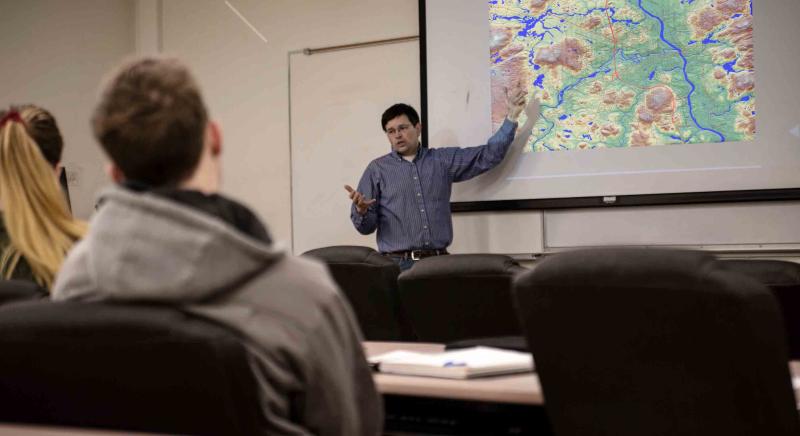 A professor pointing at a screen projection of a topographical map with students looking on from the classroom