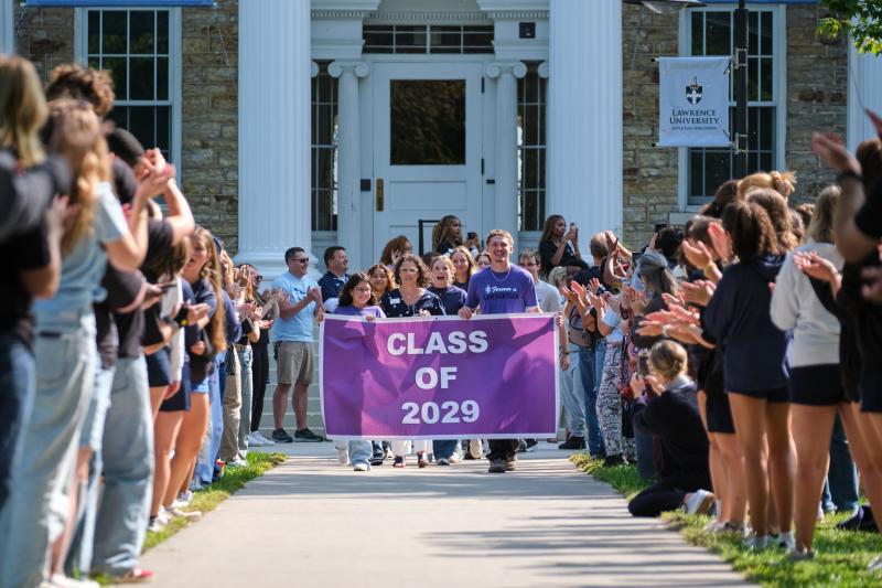 A group of students holding a purple "Class of 2029" banner walking down a path as people on both sides cheer in front of a university building.