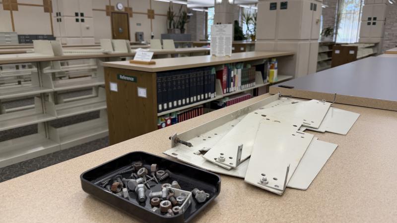 A tray of screws, nuts, and bolts and metal shelf parts on top of a library book shelf. Some filled and some empty shelves are in the background.