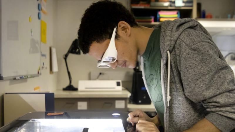 Kelvin Maestre, wearing white goggles, uses a laser cutter in the Makerspace.