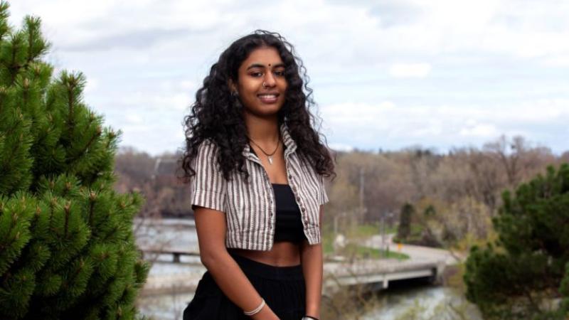Adya Kadambari, wearing a white and grey striped shirt, smiles at the camera.