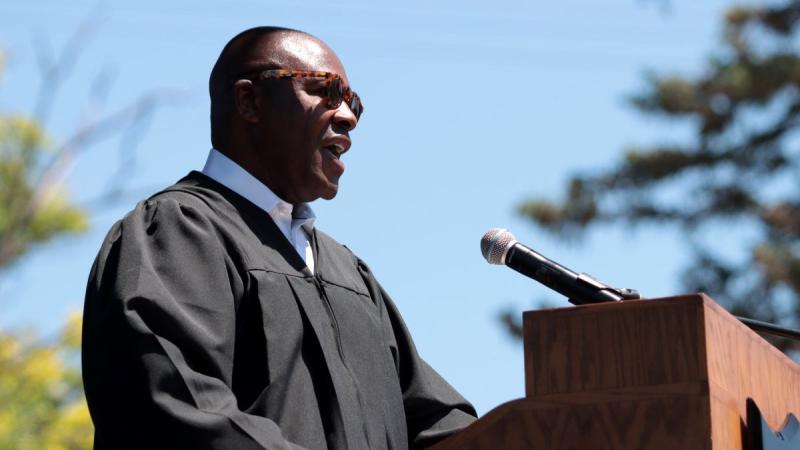Nettles behind a podium, wearing academic regalia and sunglasses
