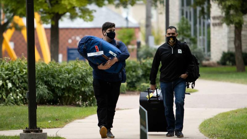 New student holds luggage, walks down campus walkway with parent pulling a suitcase