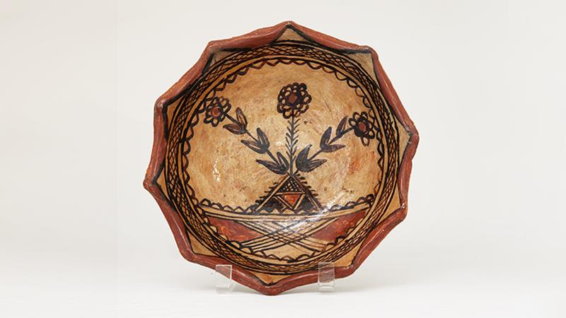 A bowl with scalloped edges and glazed in earth tones. The interior of the bowl is decorated with a floral motif surrounded by a circular geometric boarder.