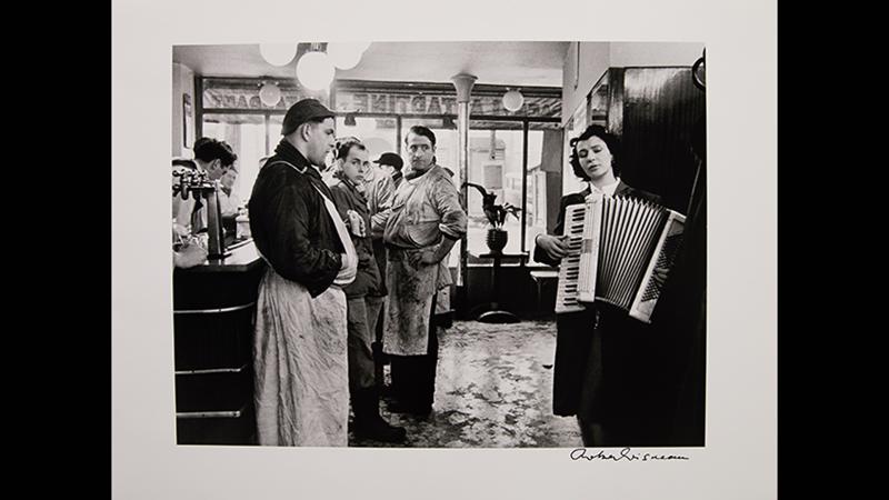 Black and white photo of a scene inside a shop. Men in workers' garbs at a counter are watching a woman play the accordion.