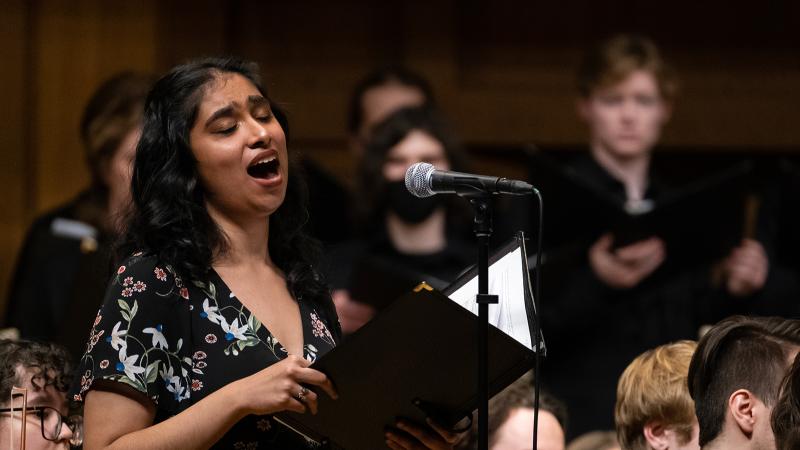 A vocalist singing a solo in performance, with an orchestra and chorus in the background.