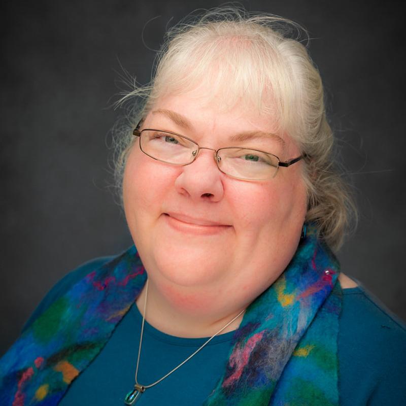 Julie is a middle-aged white woman with grey hair and glasses wearing a teal blue top and a wool nuno-felted scarf she made.