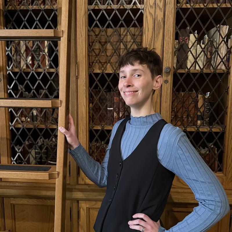 Person wearing a long-sleeved blue turlte-neck with a black vest, standing next to a ladder in a library