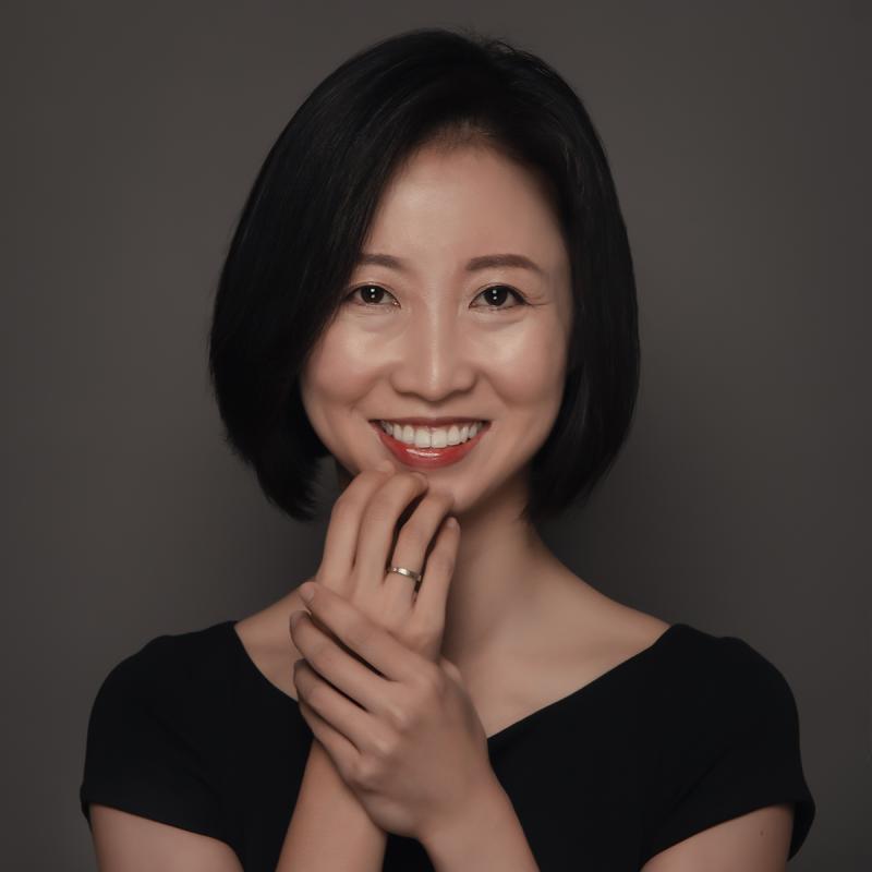 Woman with short black hair smiling, wearing a black top, and holding her hands together near her face against a plain dark background.