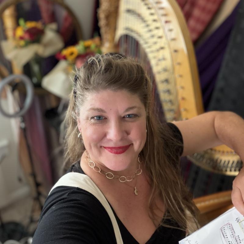 A person smiling in front of a harp while holding a sheet of music.