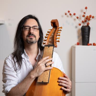 Kivie Cahn-Lipman (long-haired nerdy-looking man) holding a lirone (a 12-stringed baroque instrument). 