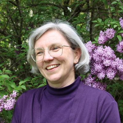 A middle-aged woman with grey hair and glasses in front of blooming lilacs