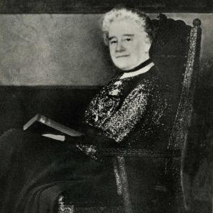 Black-and-white portrait of an older person with short, light-colored curly hair seated in a high-backed wooden chair. The person is holding an open book on their lap and faces the camera. They wear a dark, patterned outfit with a light collar.