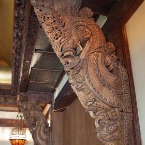Ornately carved teakwood bracket with floral and vine motifs, supporting a decorative doorway in a richly detailed interior.