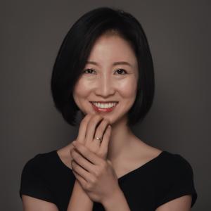 Woman with short black hair smiling, wearing a black top, and holding her hands together near her face against a plain dark background.